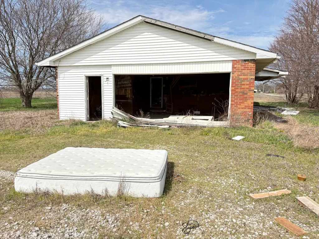 A mattress sits in the weedy, overgrown gravel driveway in front of an old garage built onto the house, with its overhead door collapsed onto the ground, and a dark shell of a house barely visible inside.